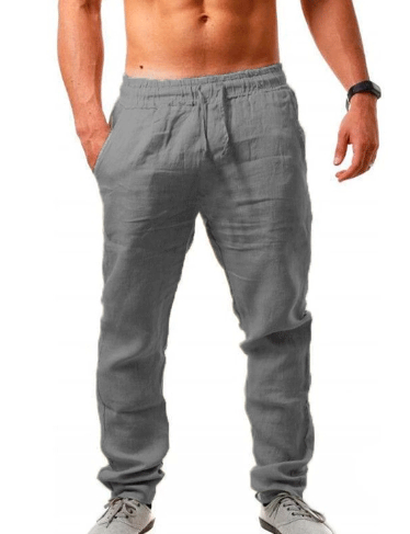 Homme portant un pantalon en lin gris, taille élastique, mains dans les poches, torse nu, montre noire, chaussures blanches. Mode décontractée masculine.