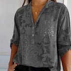 Chemisier gris à motifs floraux, manches retroussées, poche pailletée, col en V, porté par une femme. Mode élégante et décontractée.