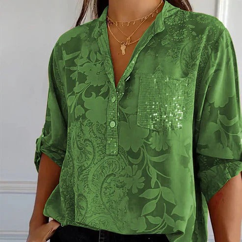 Chemisier vert à motifs floraux, manches retroussées, poche pailletée, col en V, porté avec colliers dorés. Mode élégante et tendance.