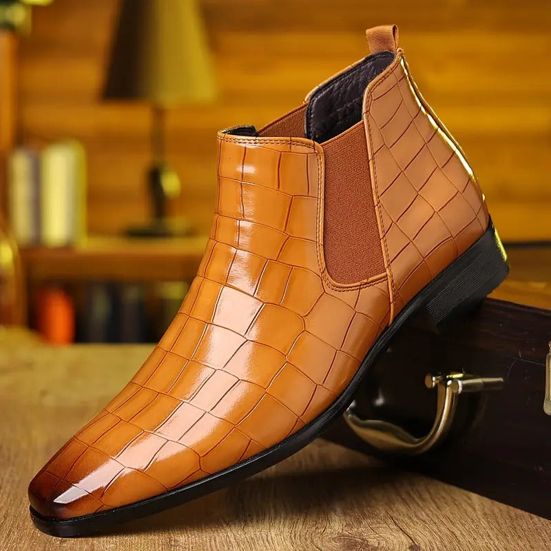 Bottine en cuir marron à motif crocodile, style Chelsea, posée sur une table en bois, avec fond flou de bibliothèque et lampe. Chaussure élégante homme.