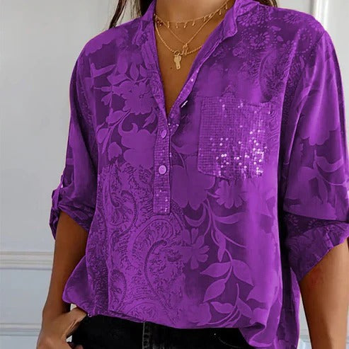 Chemisier violet à motifs floraux, manches retroussées, poche pailletée, porté par une femme avec colliers dorés. Mode féminine élégante.