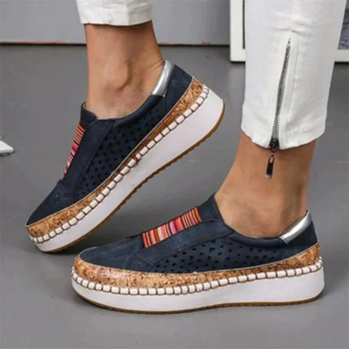 Chaussures compensées bleu marine avec semelle en liège, détails perforés et bandes colorées, portées avec un pantalon blanc zippé.