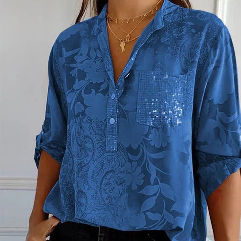 Chemisier bleu à motifs floraux, manches retroussées, poche pailletée, col en V, porté avec un collier doré. Mode féminine élégante et tendance.