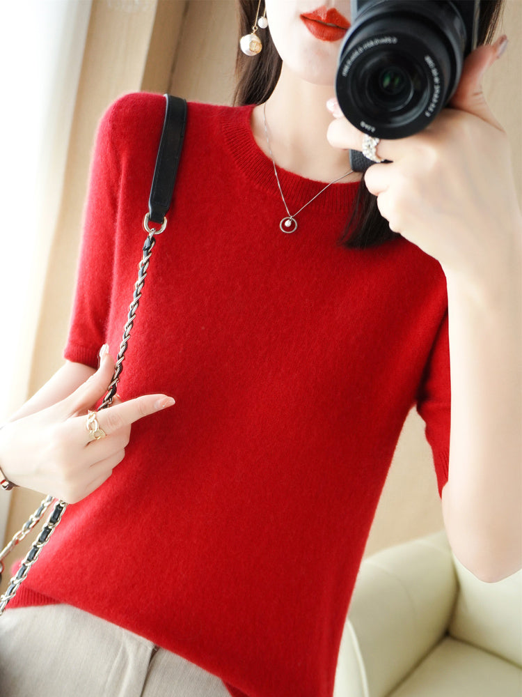 Femme en pull rouge en laine, tenant un appareil photo, portant un collier et des boucles d'oreilles dorées, intérieur lumineux.