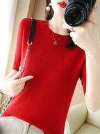 Femme en pull rouge en laine, tenant un appareil photo, portant un collier et des boucles d'oreilles dorées, intérieur lumineux.