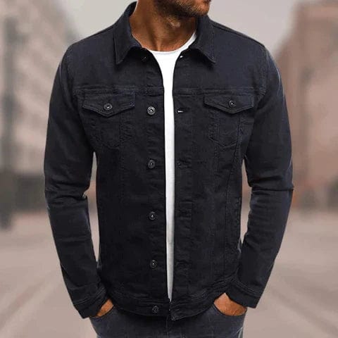 Homme portant une veste en jean noire, style décontracté, avec un t-shirt blanc en dessous, dans un environnement urbain flou. Mode masculine tendance.