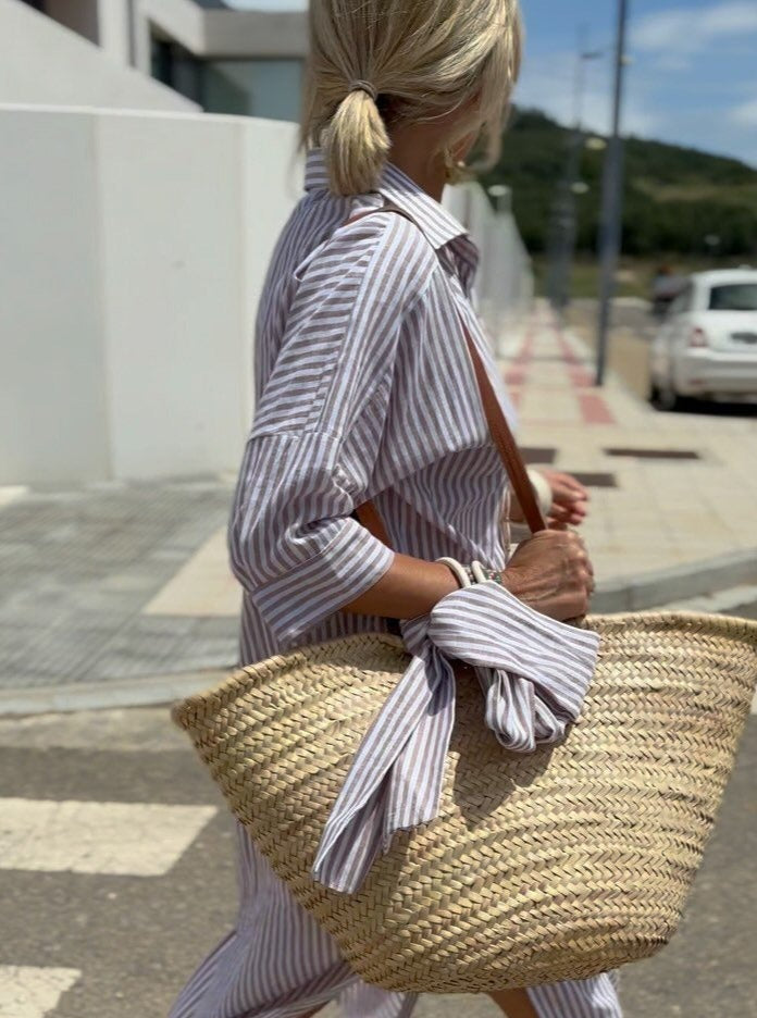 Femme en chemise rayée, portant un sac en osier sur une rue ensoleillée. Mode estivale, style décontracté, accessoires tendance.