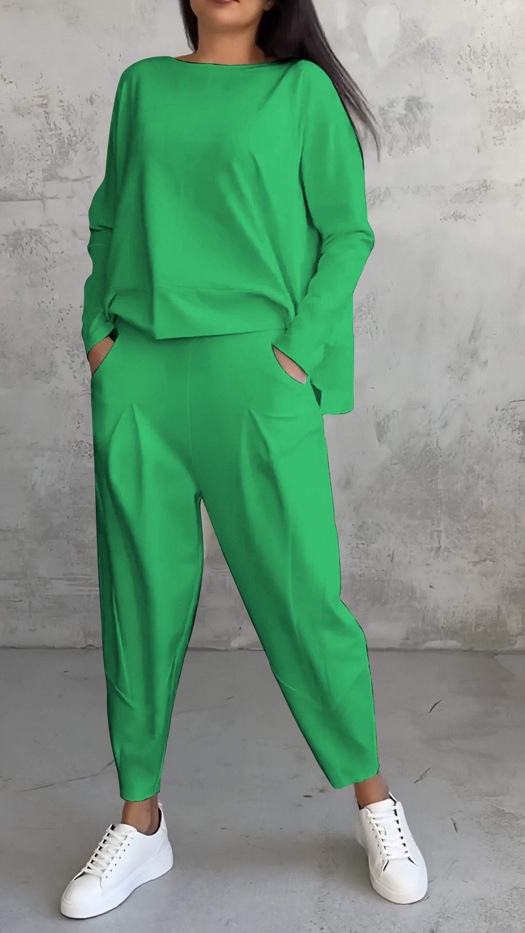 Femme en ensemble vert vif, pantalon ample et haut à manches longues, posant contre un mur texturé gris, portant des baskets blanches.