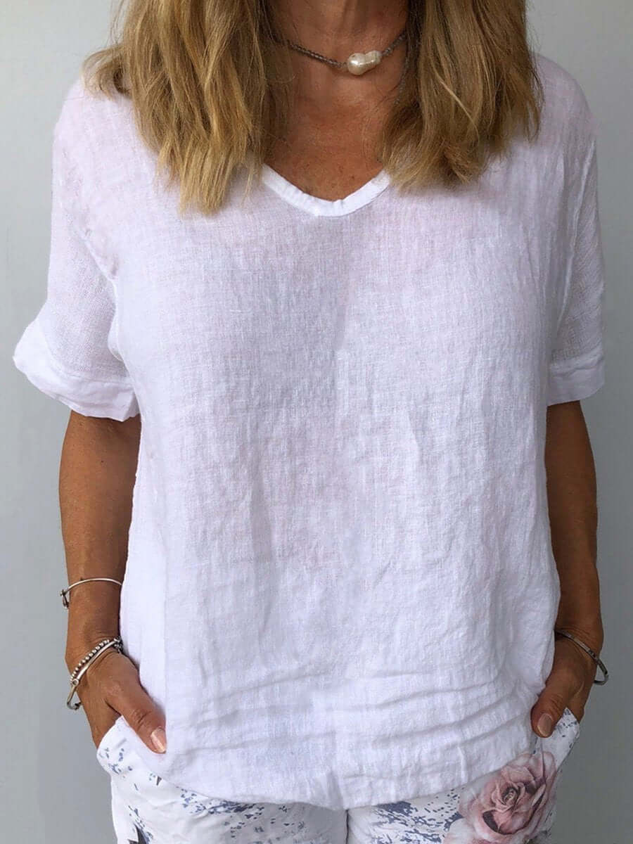 Femme portant un t-shirt blanc en lin, manches courtes, col en V, avec un collier perle, posant les mains dans les poches d'un pantalon à motifs.