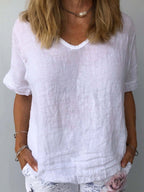 Femme portant un t-shirt blanc en lin, manches courtes, col en V, avec un collier perle, posant les mains dans les poches d'un pantalon à motifs.