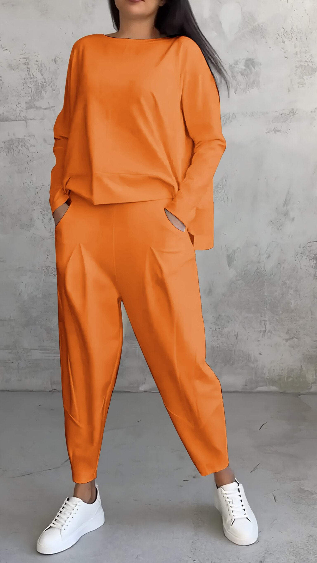 Femme portant un ensemble orange moderne avec un haut ample et un pantalon large, posant contre un mur gris, chaussures blanches. Mode décontractée élégante.