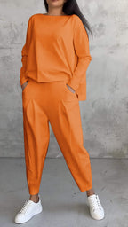 Femme portant un ensemble orange moderne avec un haut ample et un pantalon large, posant contre un mur gris, chaussures blanches. Mode décontractée élégante.