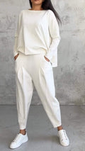 Femme en tenue décontractée blanche, portant un ensemble pull et pantalon ample, avec baskets blanches, posant devant un mur texturé gris.
