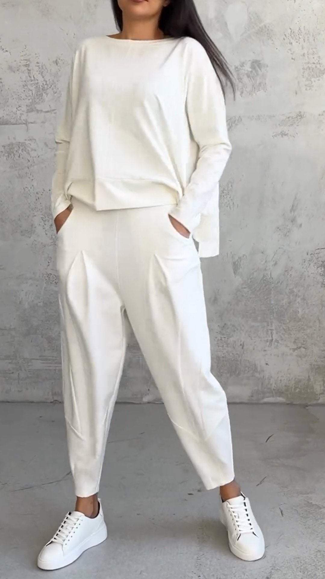 Femme en tenue décontractée blanche, portant un ensemble pull et pantalon ample, avec baskets blanches, posant devant un mur texturé gris.