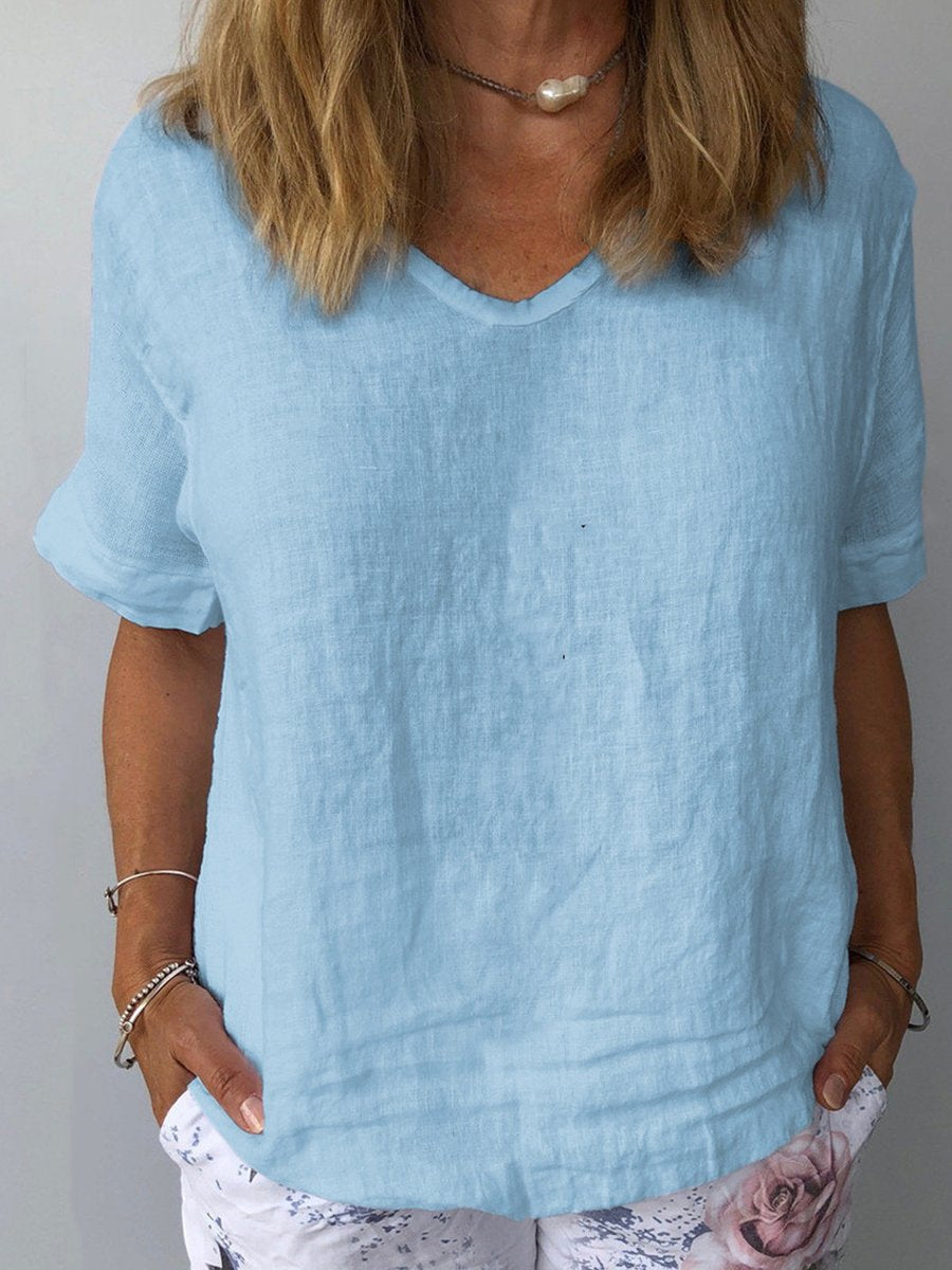 Femme portant un t-shirt en lin bleu clair avec un col en V, manches courtes, et un collier perle, sur fond neutre. Mode décontractée été.