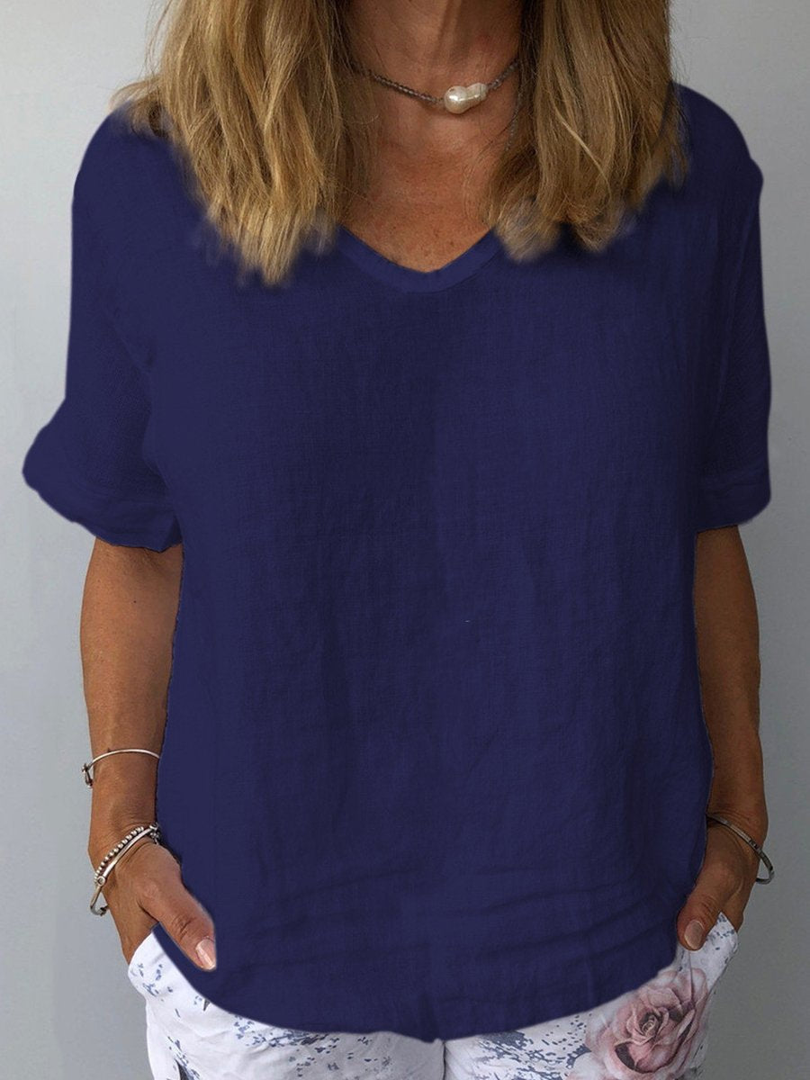 Femme portant un t-shirt bleu marine en lin, manches courtes, col en V, avec un collier perle et un pantalon blanc à motif floral. Mode décontractée élégante.