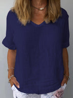 Femme portant un t-shirt bleu marine en lin, manches courtes, col en V, avec un collier perle et un pantalon blanc à motif floral. Mode décontractée élégante.