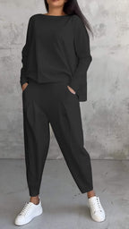 Femme en tenue décontractée noire, pantalon ample et pull, posant contre un mur texturé gris, portant des baskets blanches. Mode urbaine chic.