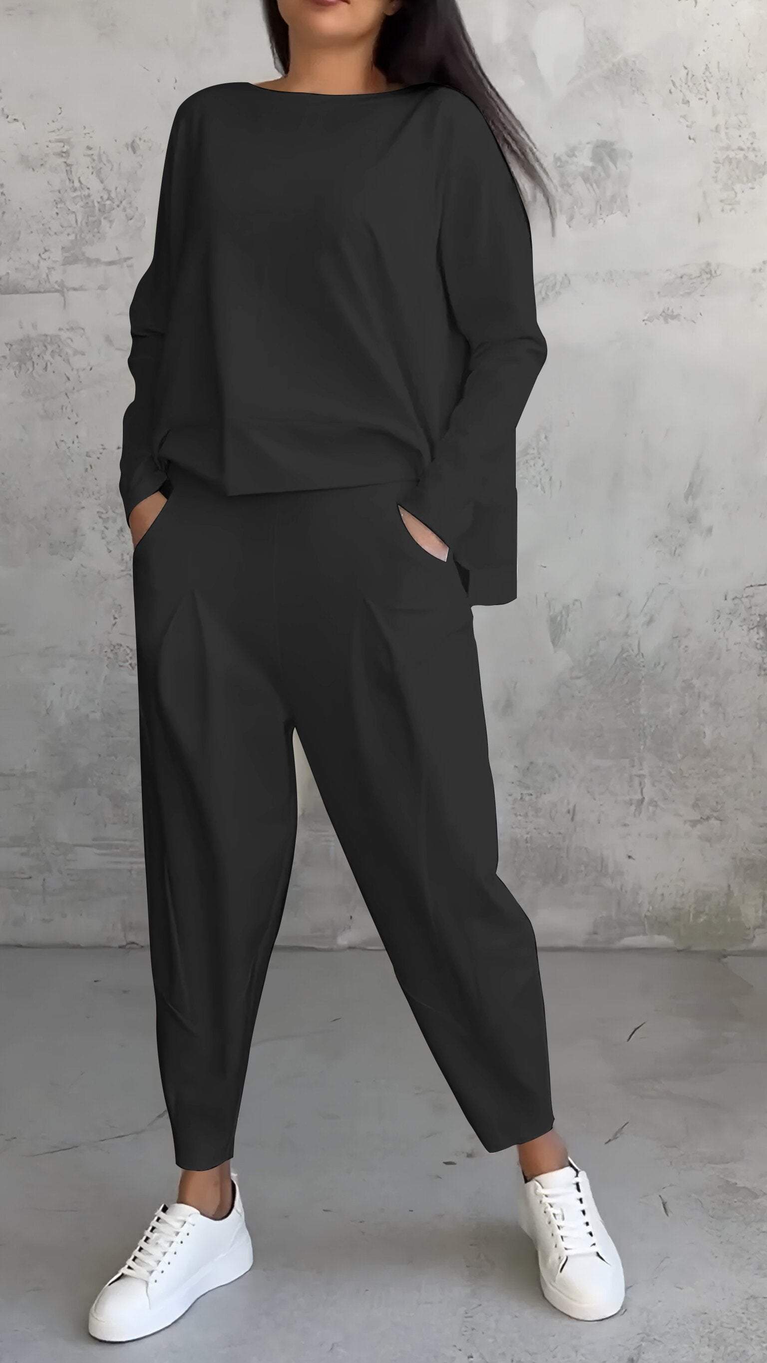 Femme en tenue décontractée noire, pantalon ample et pull, posant contre un mur texturé gris, portant des baskets blanches. Mode urbaine chic.