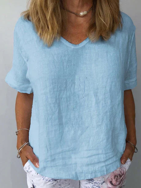 Femme portant un t-shirt bleu clair en lin, manches courtes, col en V, avec un collier perle, et un pantalon blanc à motif floral.