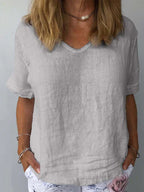 Femme portant un t-shirt en lin gris clair, col en V, manches courtes, avec un collier perle, et un pantalon blanc à motif floral. Mode décontractée élégante.