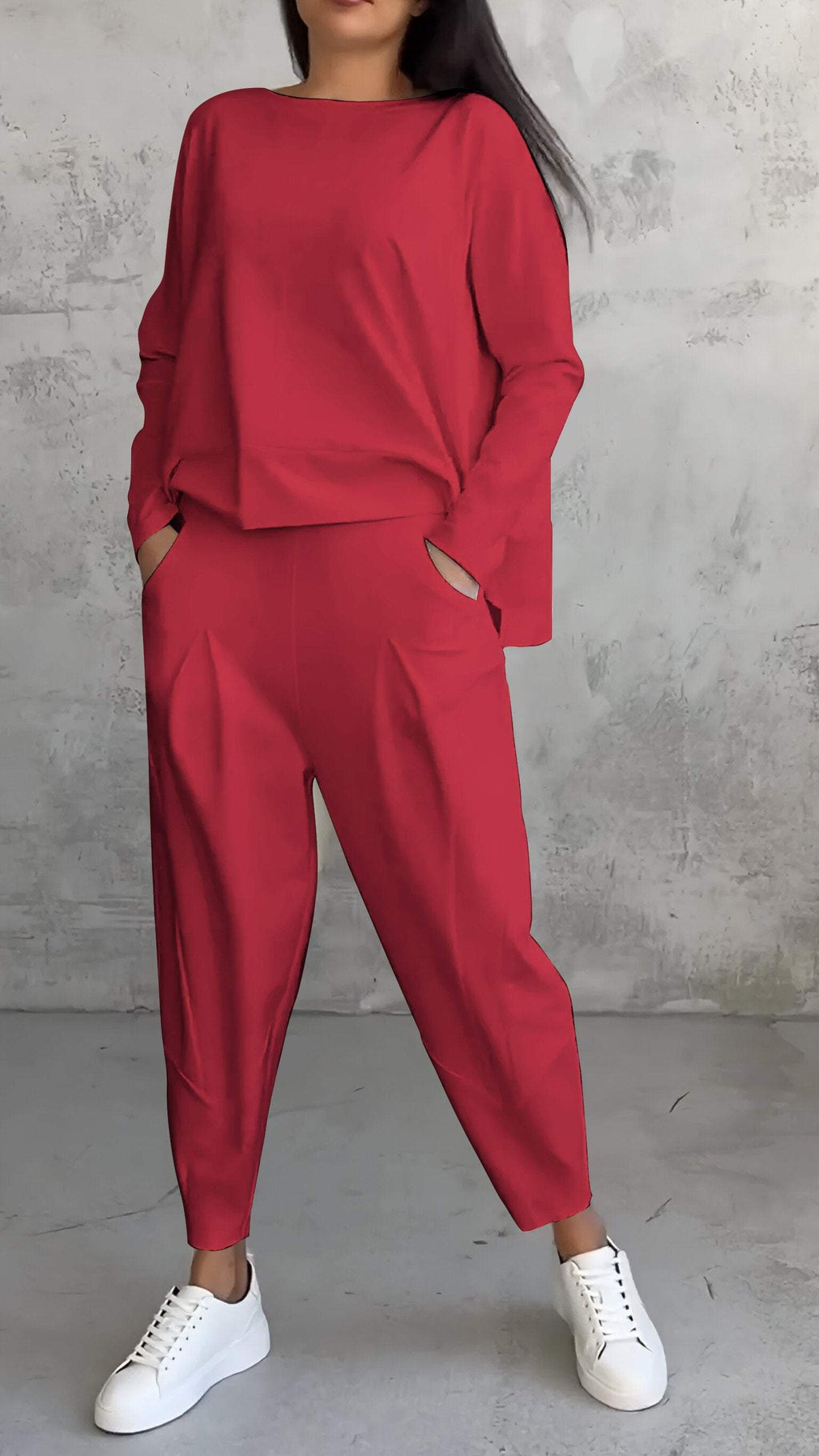 Femme portant un ensemble rouge élégant avec un haut ample et un pantalon taille haute, posant contre un mur texturé gris, chaussures blanches.