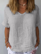 Femme portant un t-shirt en lin gris à col en V, manches courtes, avec un collier perle et un pantalon blanc à motif floral. Mode décontractée élégante.