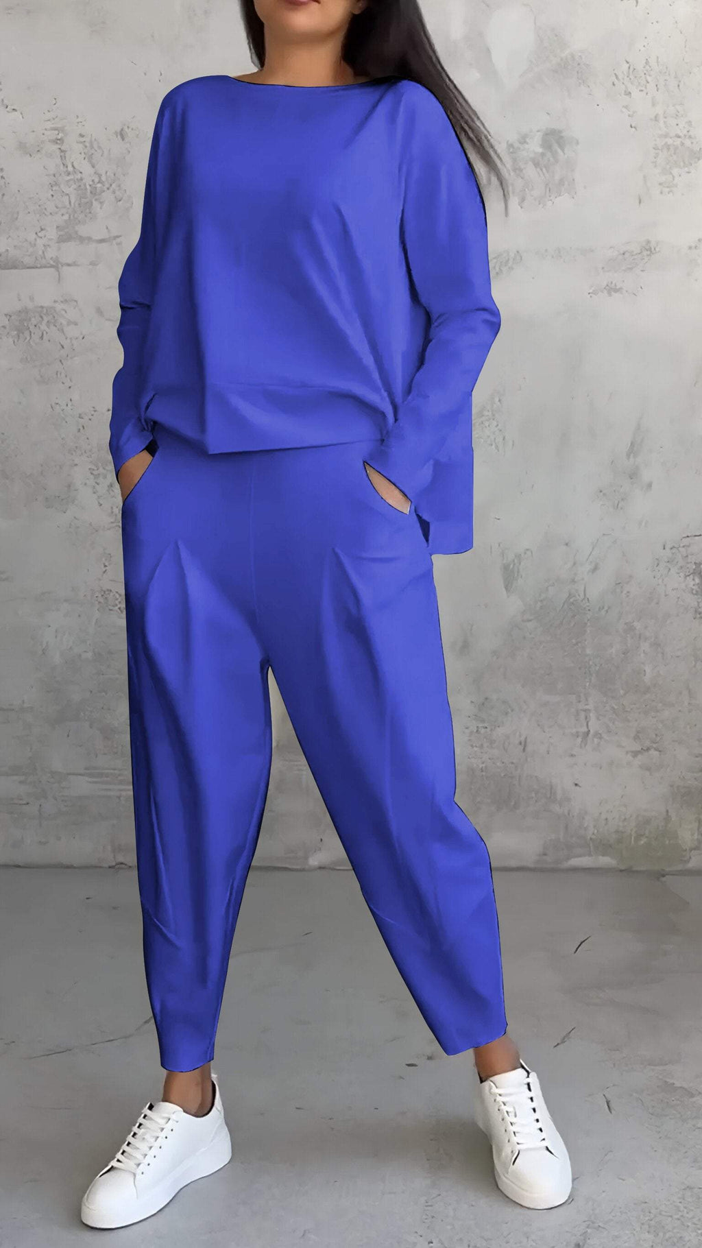 Femme en ensemble bleu électrique, pantalon ample et haut à manches longues, posant contre un mur texturé gris, portant des baskets blanches.