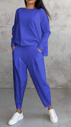 Femme en ensemble bleu électrique, pantalon ample et haut à manches longues, posant contre un mur texturé gris, portant des baskets blanches.