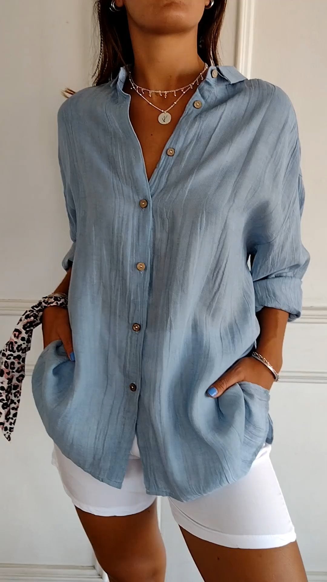 Femme portant une chemise bleue en lin avec boutons en bois, manches retroussées, associée à un short blanc. Style décontracté et élégant.