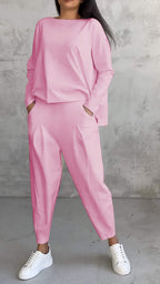Femme portant un ensemble rose ample avec des baskets blanches, posant devant un mur texturé gris. Mode décontractée, style moderne.