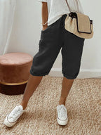Femme portant un short noir décontracté, t-shirt blanc, baskets blanches et sac à main en paille, debout sur un tapis beige. Mode estivale chic.