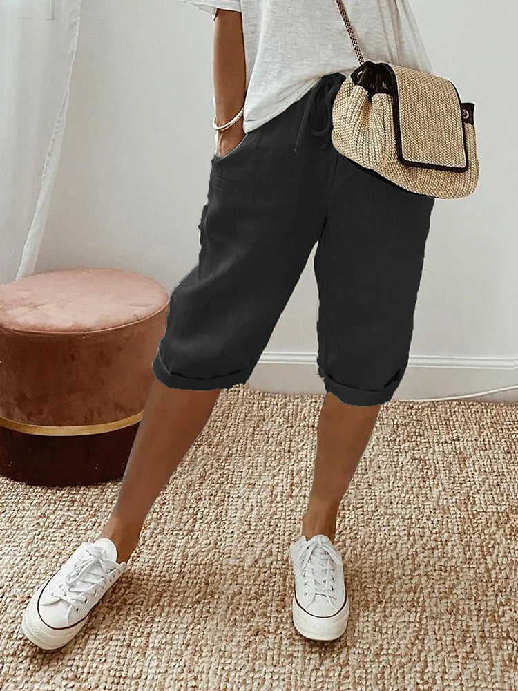 Femme en short noir décontracté, t-shirt blanc, sac à main en paille, baskets blanches, posant sur un tapis beige. Mode estivale chic et confortable.