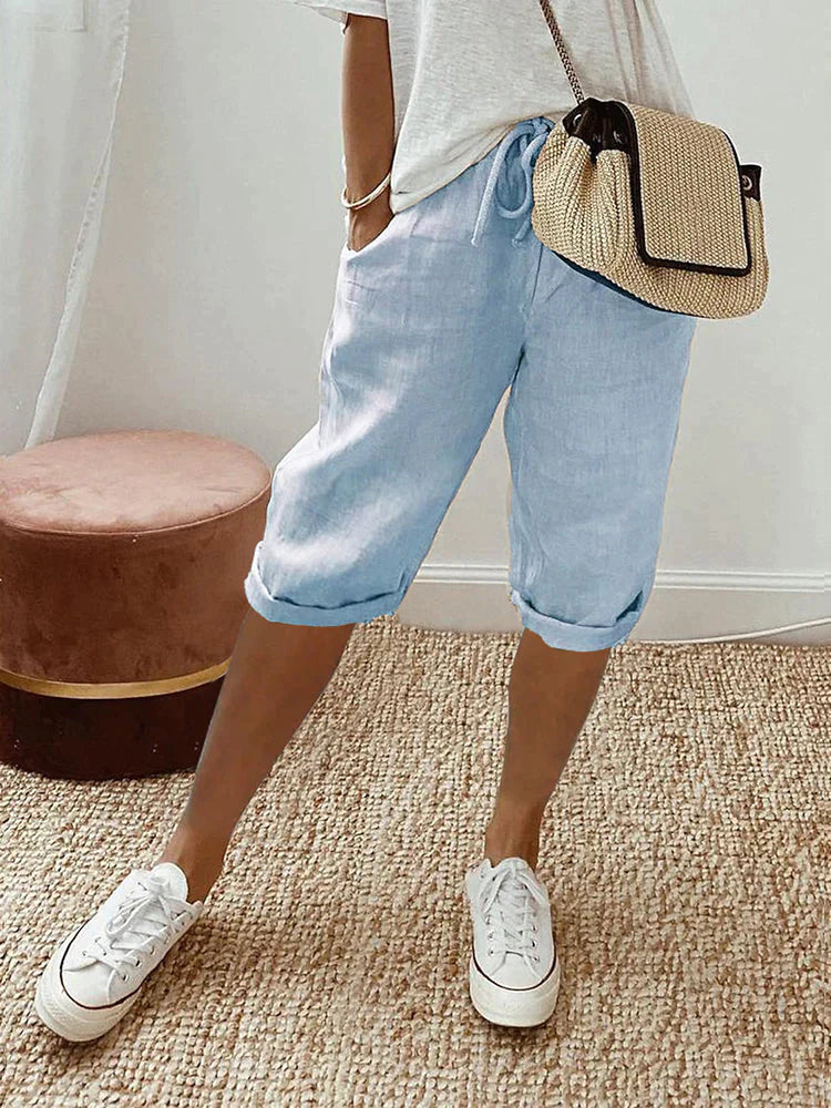 Femme en short bleu clair et t-shirt blanc, portant un sac à main en paille, baskets blanches, sur tapis beige. Mode décontractée estivale.