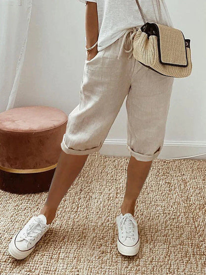 Femme portant un short en lin beige, t-shirt blanc, baskets blanches et sac à main en paille, posant dans une pièce moderne.