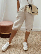 Personne portant un short en lin beige, t-shirt blanc, baskets blanches et sac à main en paille, debout sur un tapis en jute. Mode décontractée estivale.