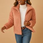 Femme souriante portant une veste en fausse fourrure rose, pull blanc à col roulé, jeans bleu, fond beige. Mode automne-hiver, style élégant.