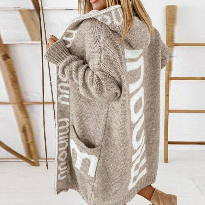 Femme portant un long cardigan beige à capuche avec motifs blancs, style décontracté, mode automne-hiver, fond échelle en bois.