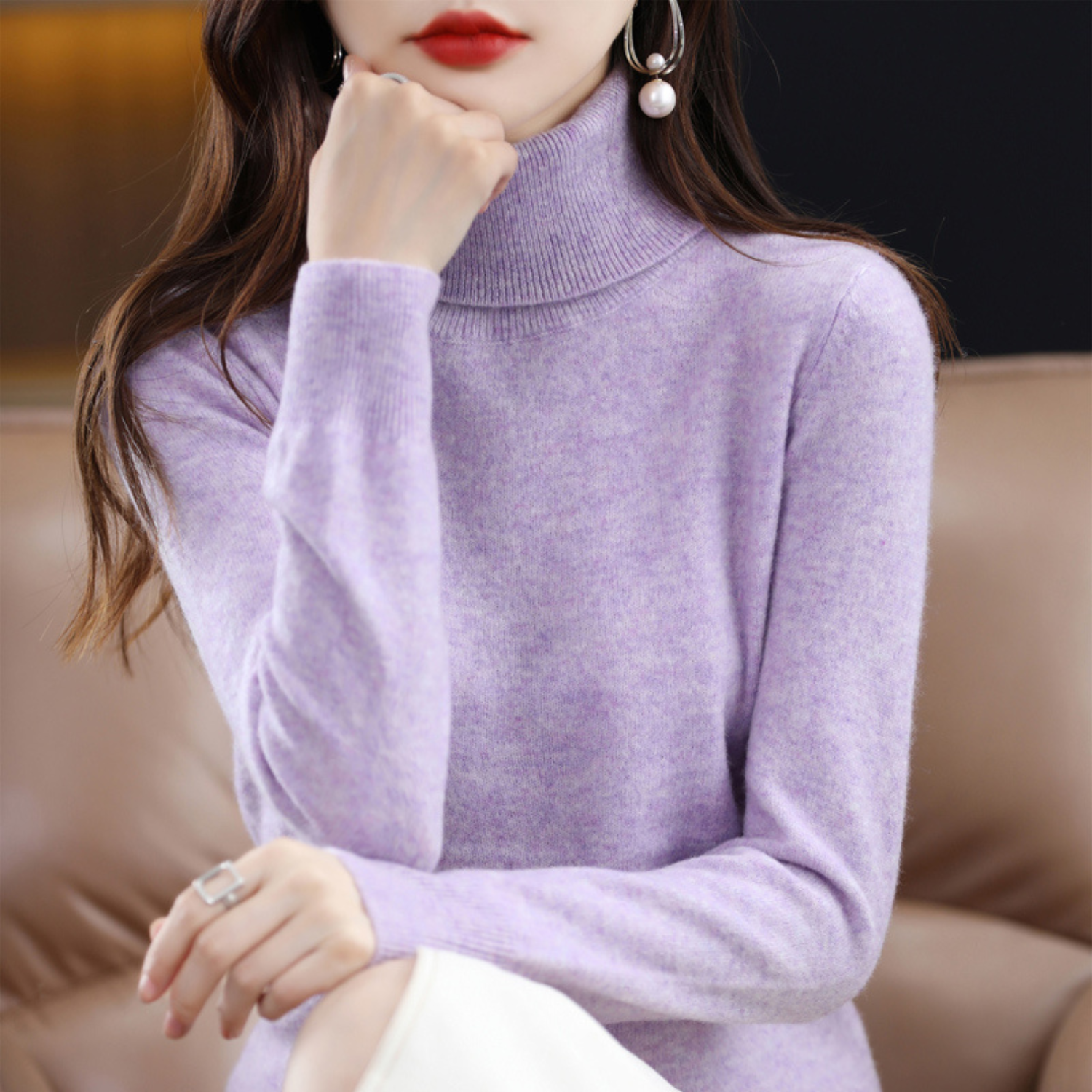 Femme portant un pull col roulé violet, assise sur un canapé beige, avec des boucles d'oreilles perle et une bague argentée. Mode élégante automne.