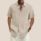 Homme portant une chemise en lin beige à manches courtes, col ouvert, avec un pantalon blanc. Mode estivale élégante et décontractée.