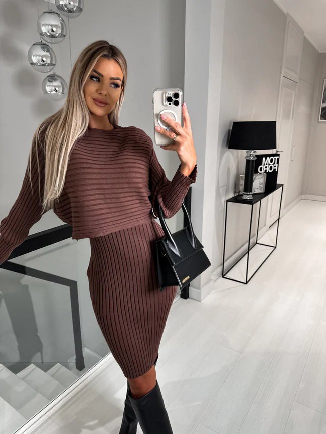 Femme prenant un selfie, portant une robe pull marron côtelée, sac à main noir, dans un intérieur moderne avec lampes suspendues et lampe noire.