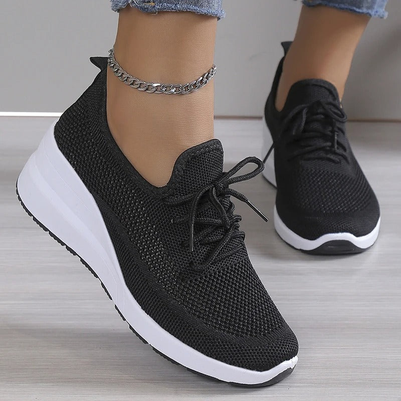 Chaussures de sport noires pour femme avec semelle blanche, portées avec un bracelet de cheville argenté. Sneakers tendance et confortables.