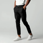 Homme portant un pantalon noir ajusté et des baskets blanches, debout sur fond gris. Mode masculine élégante et décontractée.
