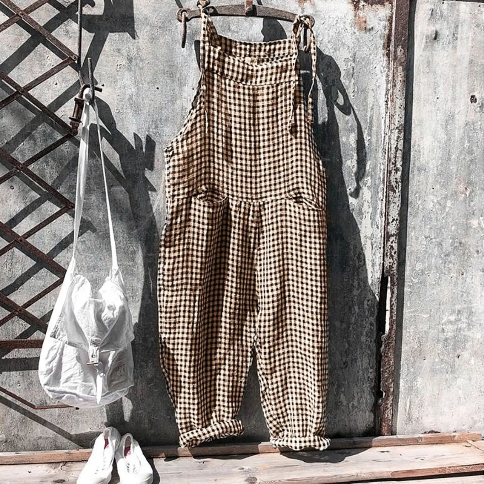 Salopette à carreaux marron suspendue sur un mur en métal, accompagnée d'un sac blanc et de chaussures blanches. Mode vintage, style décontracté.