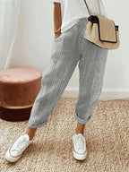 Femme portant un pantalon rayé gris et blanc, t-shirt blanc, baskets blanches, sac en osier, posant sur un tapis beige. Mode décontractée chic.