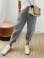 Femme portant un pantalon rayé bleu et blanc, t-shirt blanc, baskets blanches, sac en osier, debout sur un tapis beige. Mode décontractée chic.