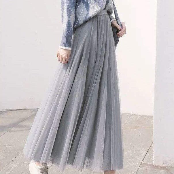Femme portant une jupe longue plissée grise, associée à un pull bleu à motifs losanges, style élégant et décontracté, mode automne-hiver.