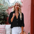 Jeune femme blonde portant une chemise noire et un short blanc, posant sur une terrasse avec des plantes et des coussins, ambiance estivale.