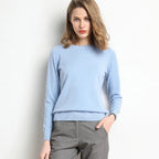 Femme portant un pull bleu clair en cachemire, posant contre un mur blanc. Mode élégante, tenue automne-hiver, style décontracté chic.