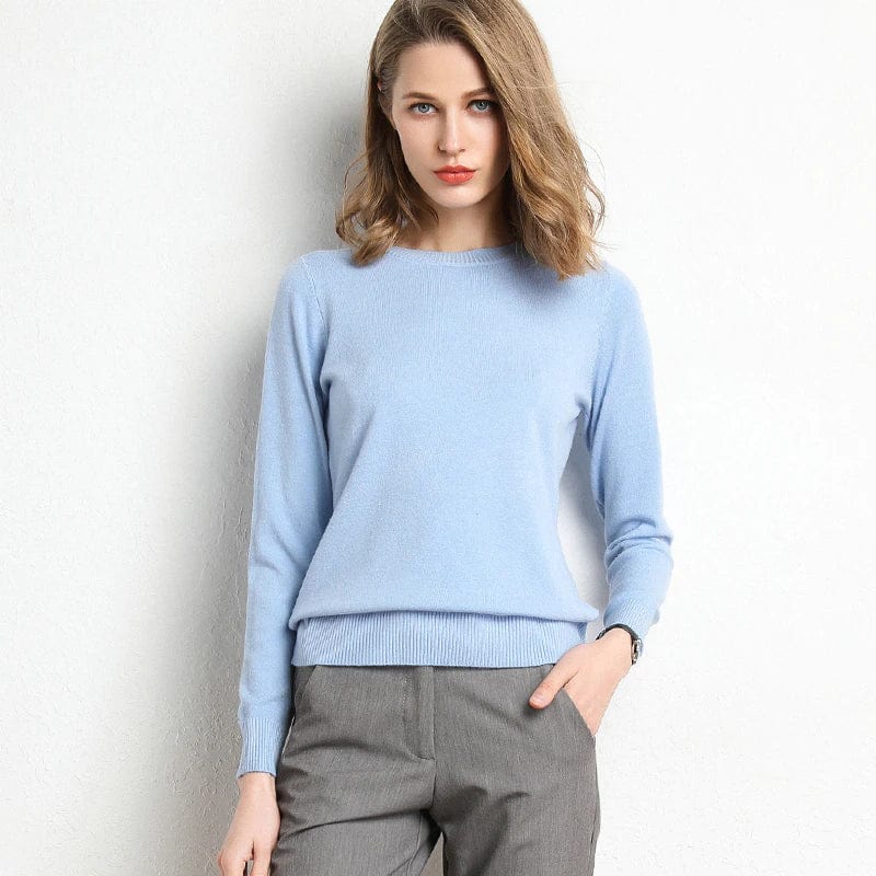 Femme portant un pull bleu clair en cachemire, posant contre un mur blanc. Mode élégante, tenue automne-hiver, style décontracté chic.